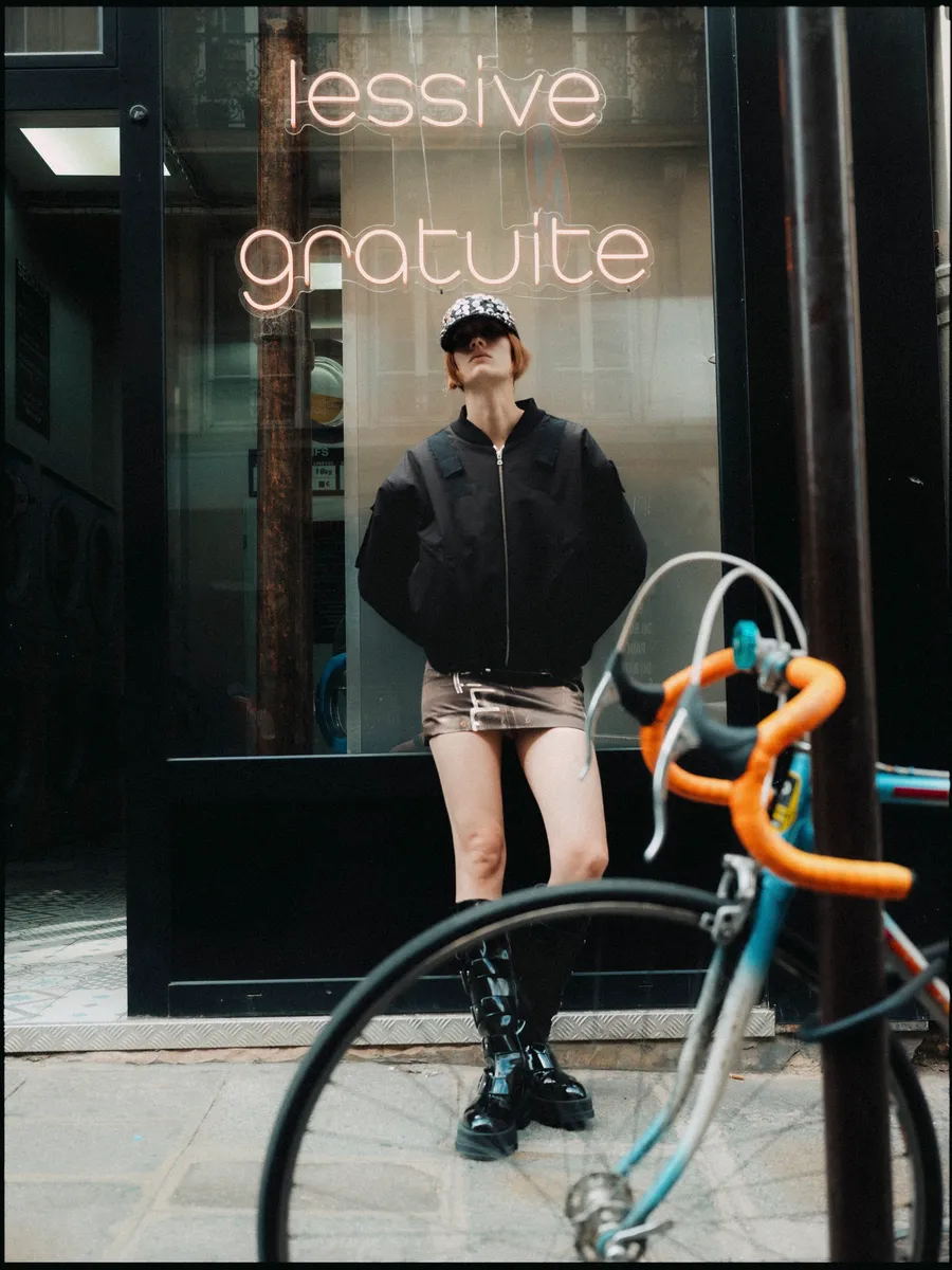 Femme en bomber noir, mini-jupe et bottes vernies devant une laverie à néon « lessive gratuite », vélo au premier plan