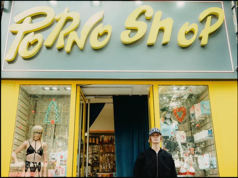 Femme en veste noire et casquette à motifs devant une devanture jaune surmontée d'une enseigne « Porno Shop »
