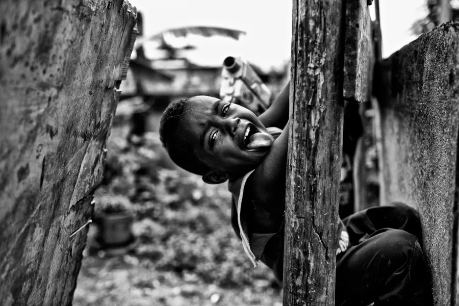 Enfant riant, accroupi entre deux palissades de bois, ruelle de favela en arrière-plan, noir et blanc