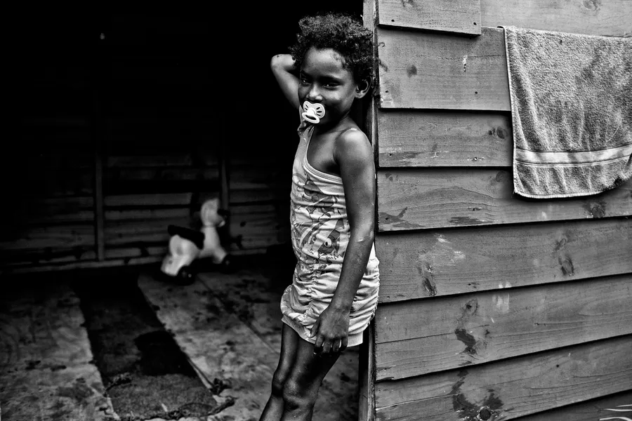Fillette à la tétine adossée à une baraque en planches, serviette suspendue, noir et blanc