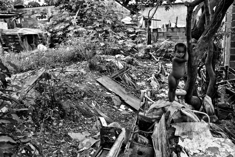 Jeune enfant nu debout contre un arbre dans un terrain jonché de gravats, noir et blanc