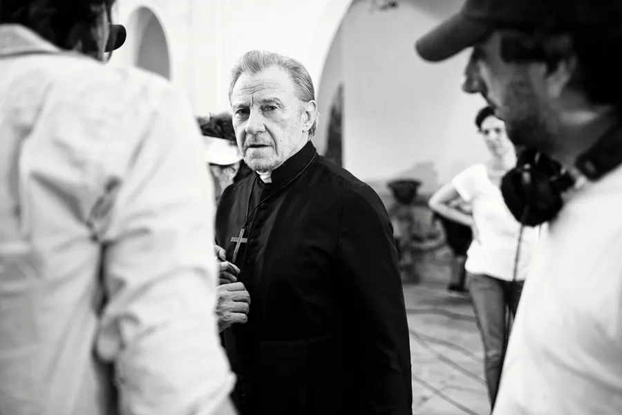 Homme en soutane noire avec croix pectorale entouré d'une équipe de tournage, noir et blanc