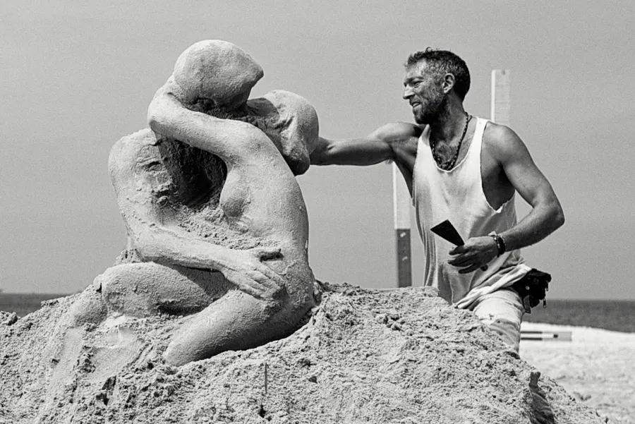 Sculpteur en débardeur façonnant une sculpture de sable de deux personnages enlacés sur une plage, noir et blanc