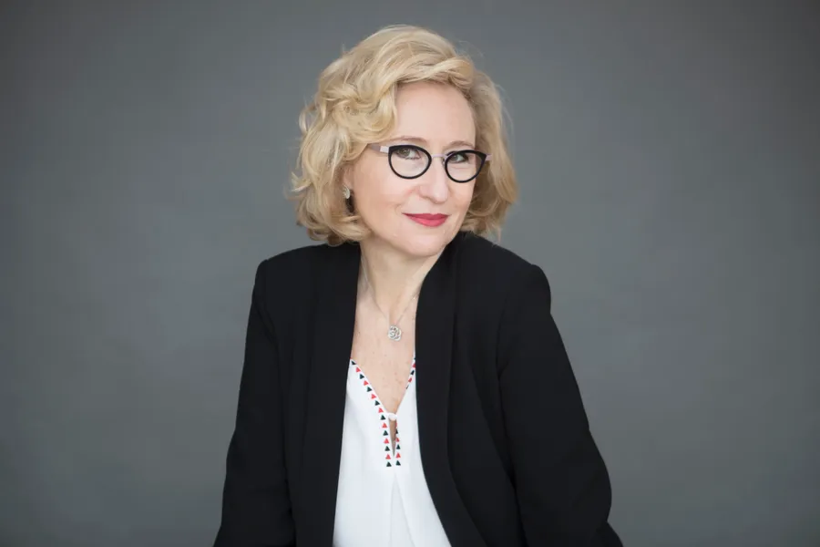 Femme blonde à lunettes en veste noire et blouse blanche brodée, fond gris uni