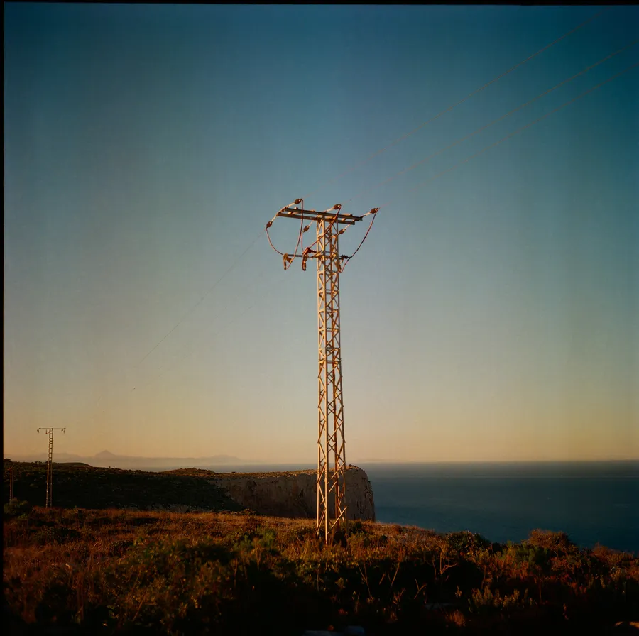 Pylône électrique dressé sur une falaise au bord de la mer, lumière rasante de fin de journée