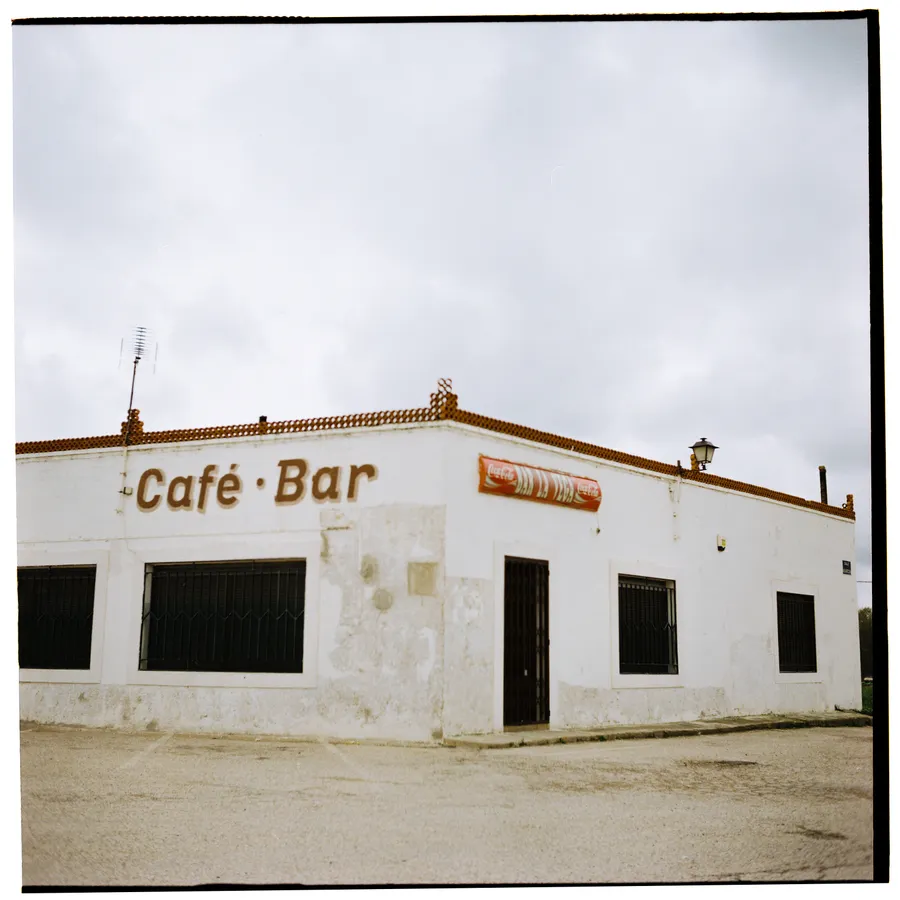 Bâtiment blanc fermé portant l'enseigne peinte « Café · Bar » au bord d'une route déserte