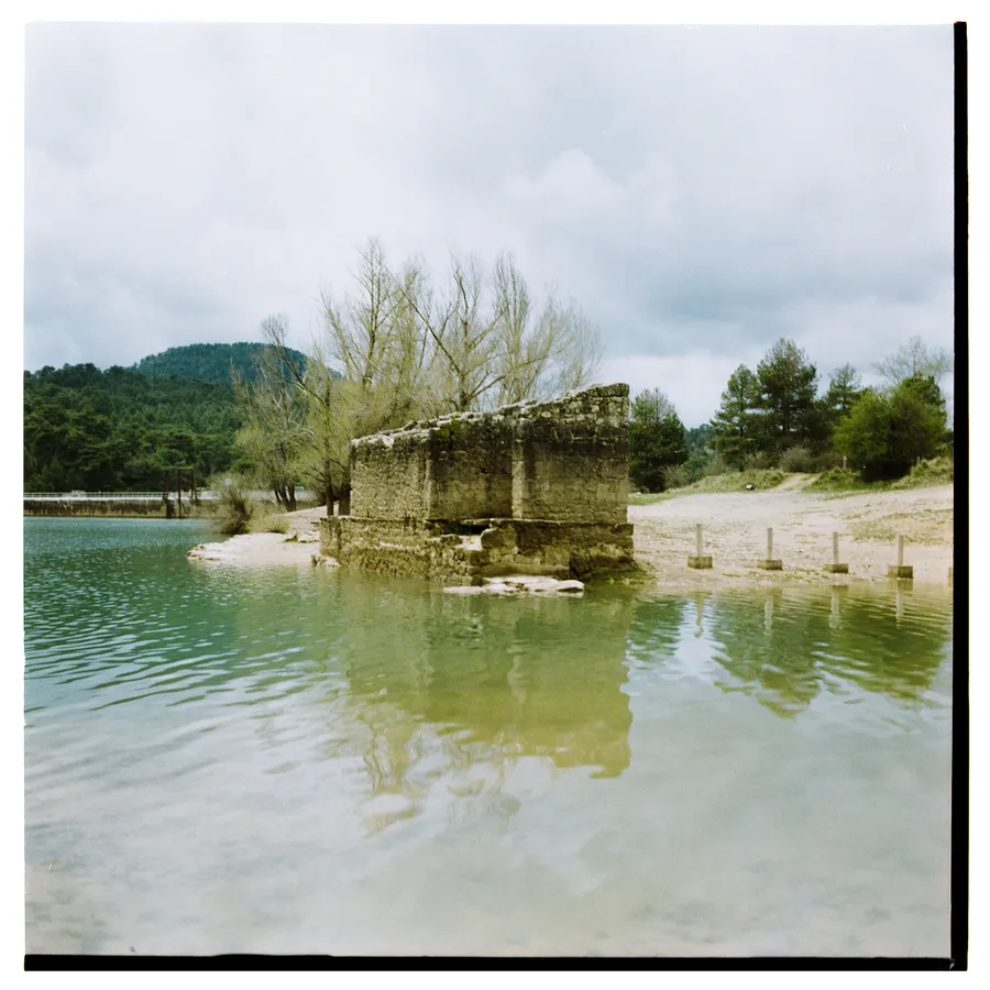 Ruine de pierre les pieds dans l'eau d'un lac, rive sableuse et collines boisées derrière