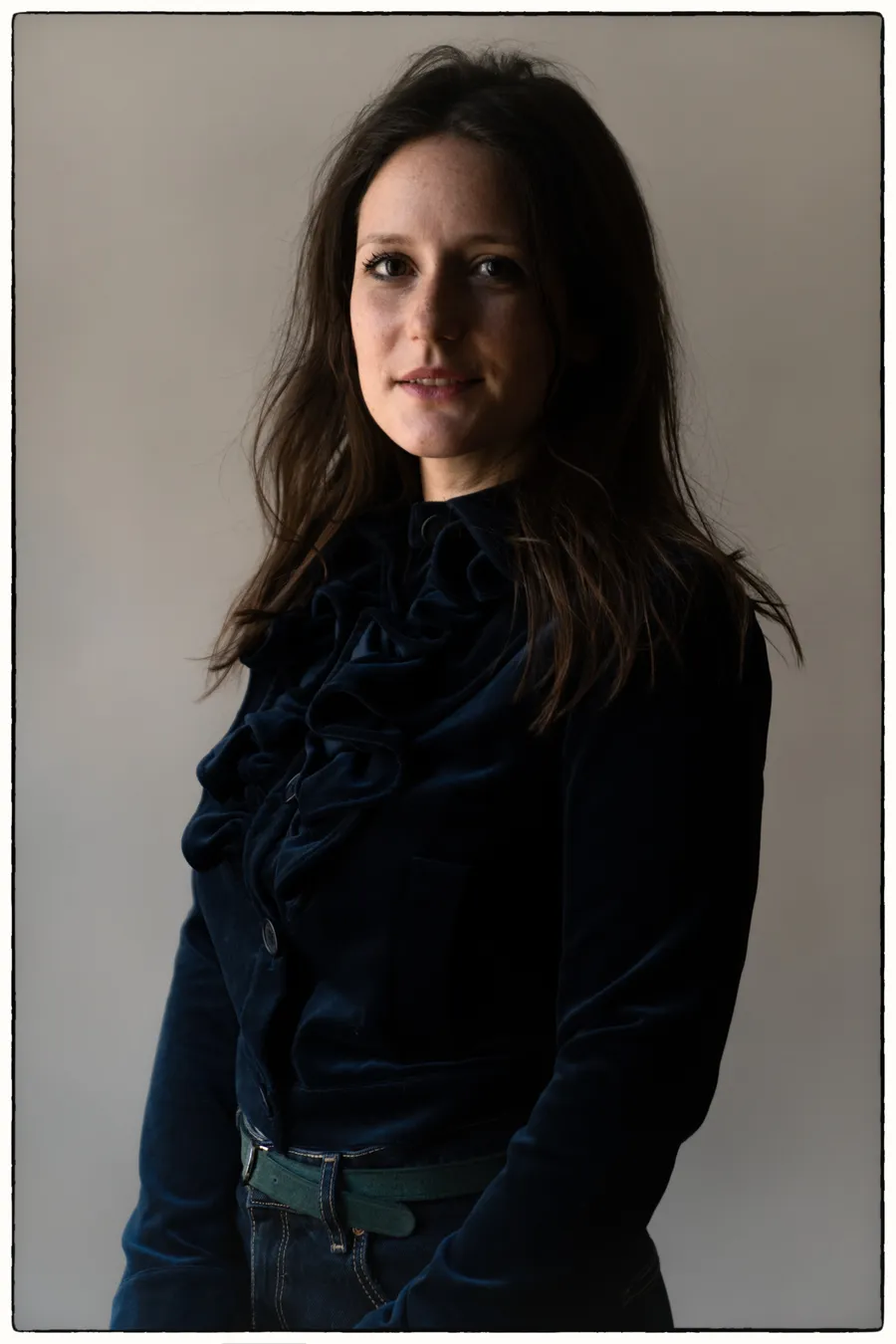 Femme brune en veste de velours foncé à jabot, debout devant un fond gris