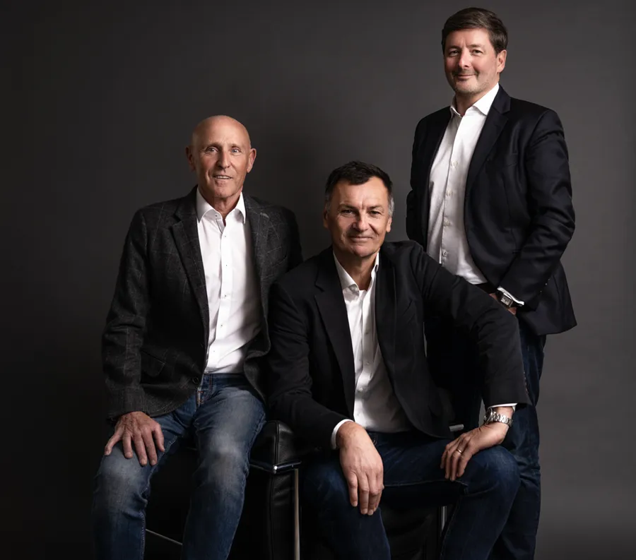 Trois hommes en veste et chemise blanche posant ensemble devant un fond gris foncé
