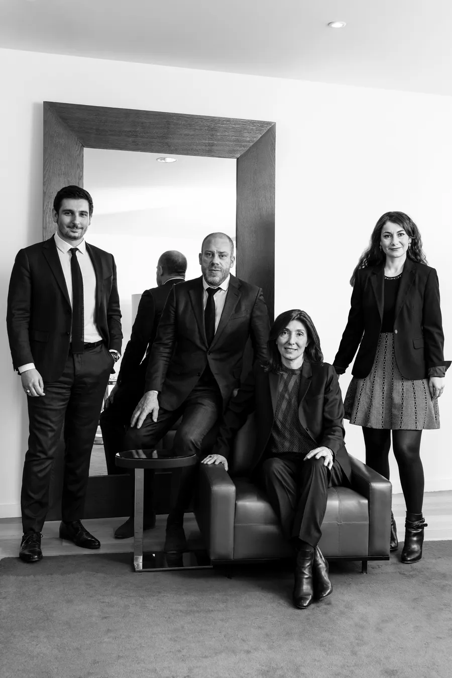 Quatre personnes en tenue de ville posant devant un grand miroir et un fauteuil, noir et blanc