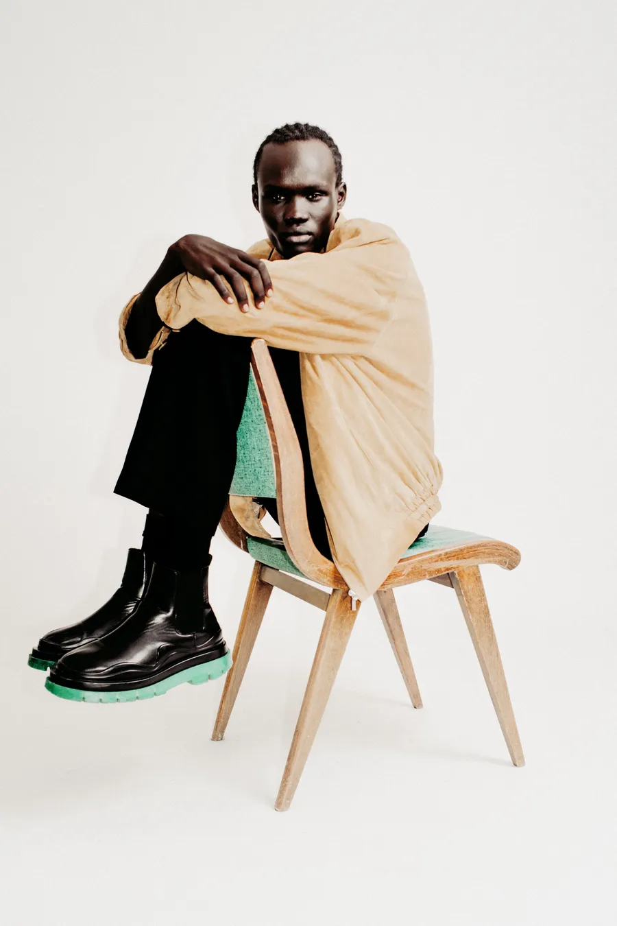 Homme assis sur une chaise en bois, genoux repliés, veste beige ample et bottines noires à semelle verte