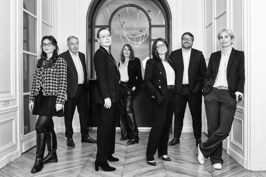 Sept personnes d'un cabinet posant dans un couloir haussmannien à parquet, noir et blanc