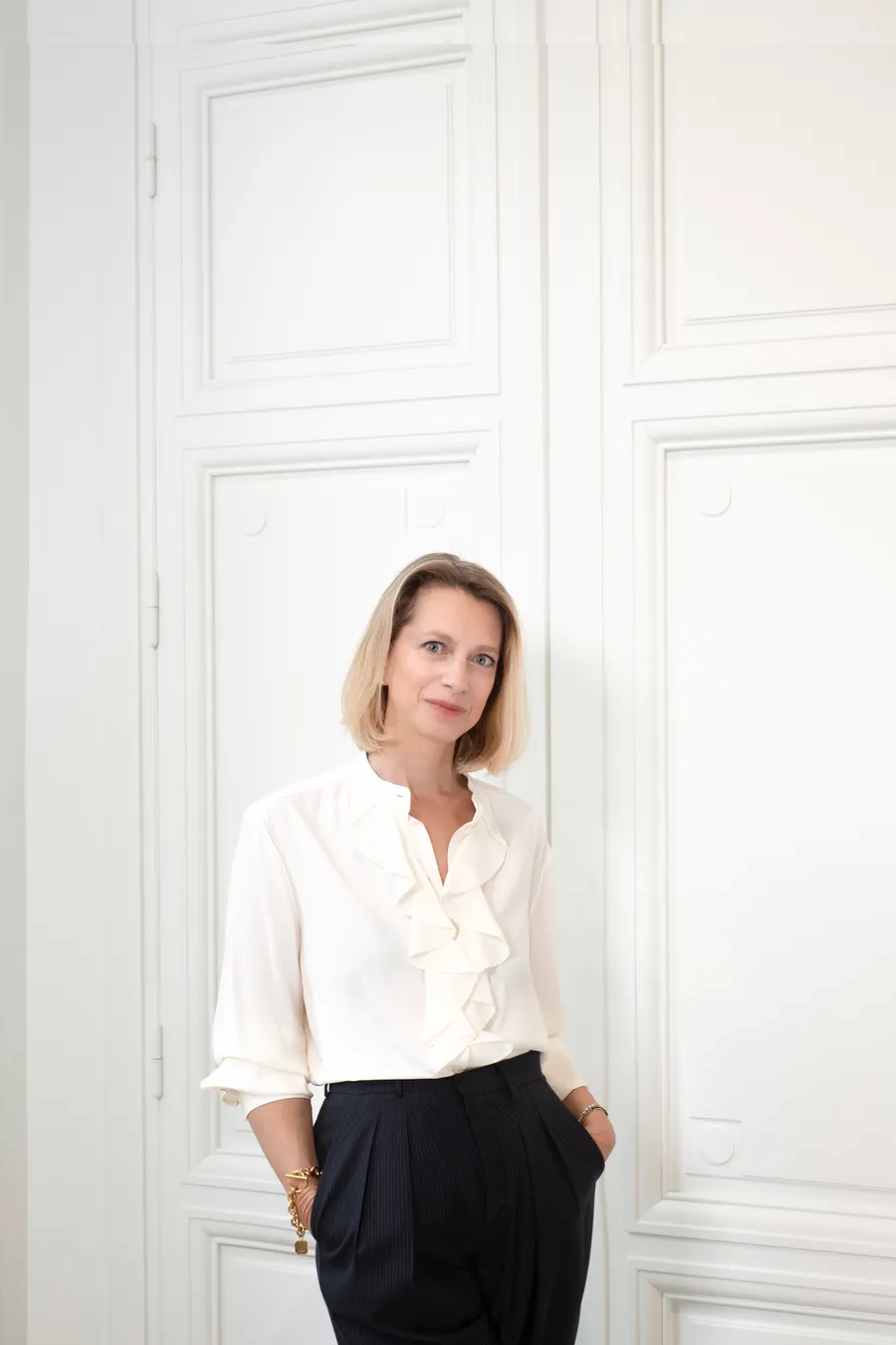 Femme blonde en chemisier blanc à jabot et pantalon foncé, devant des boiseries blanches