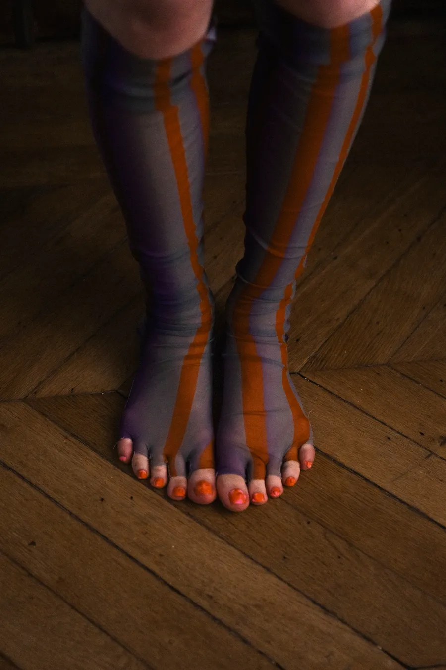 Pieds nus aux ongles orange dans des collants rayés gris et orange, sur un parquet en chevrons