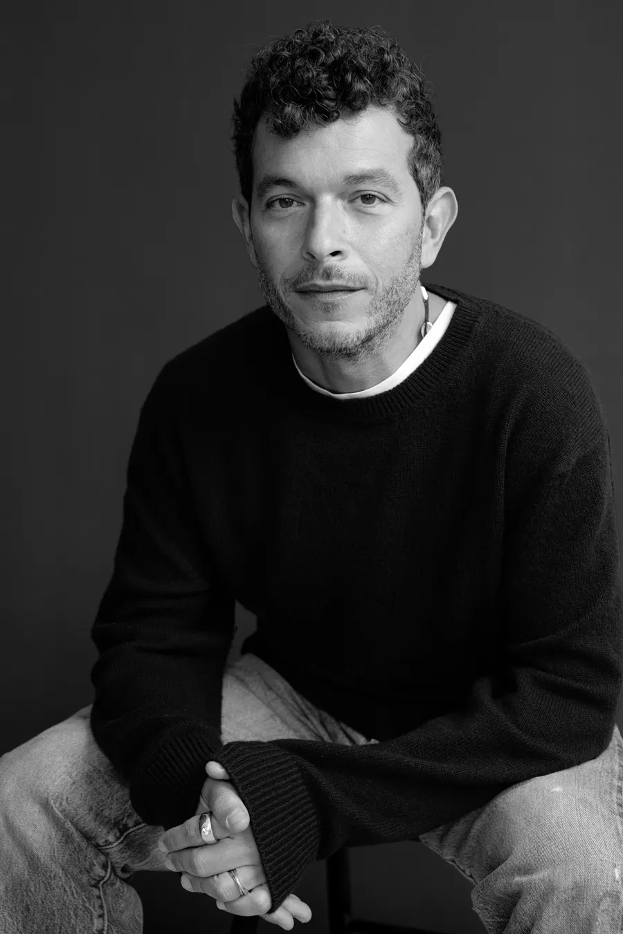 Homme aux cheveux bouclés en pull noir assis, mains jointes, fond gris, noir et blanc