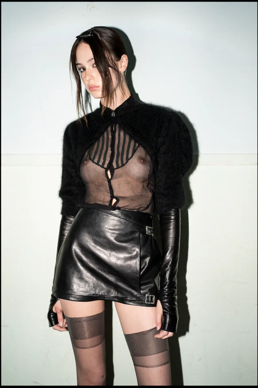 Mannequin en boléro noir duveteux, chemise transparente et jupe en cuir, gants longs et bas, contre un mur clair