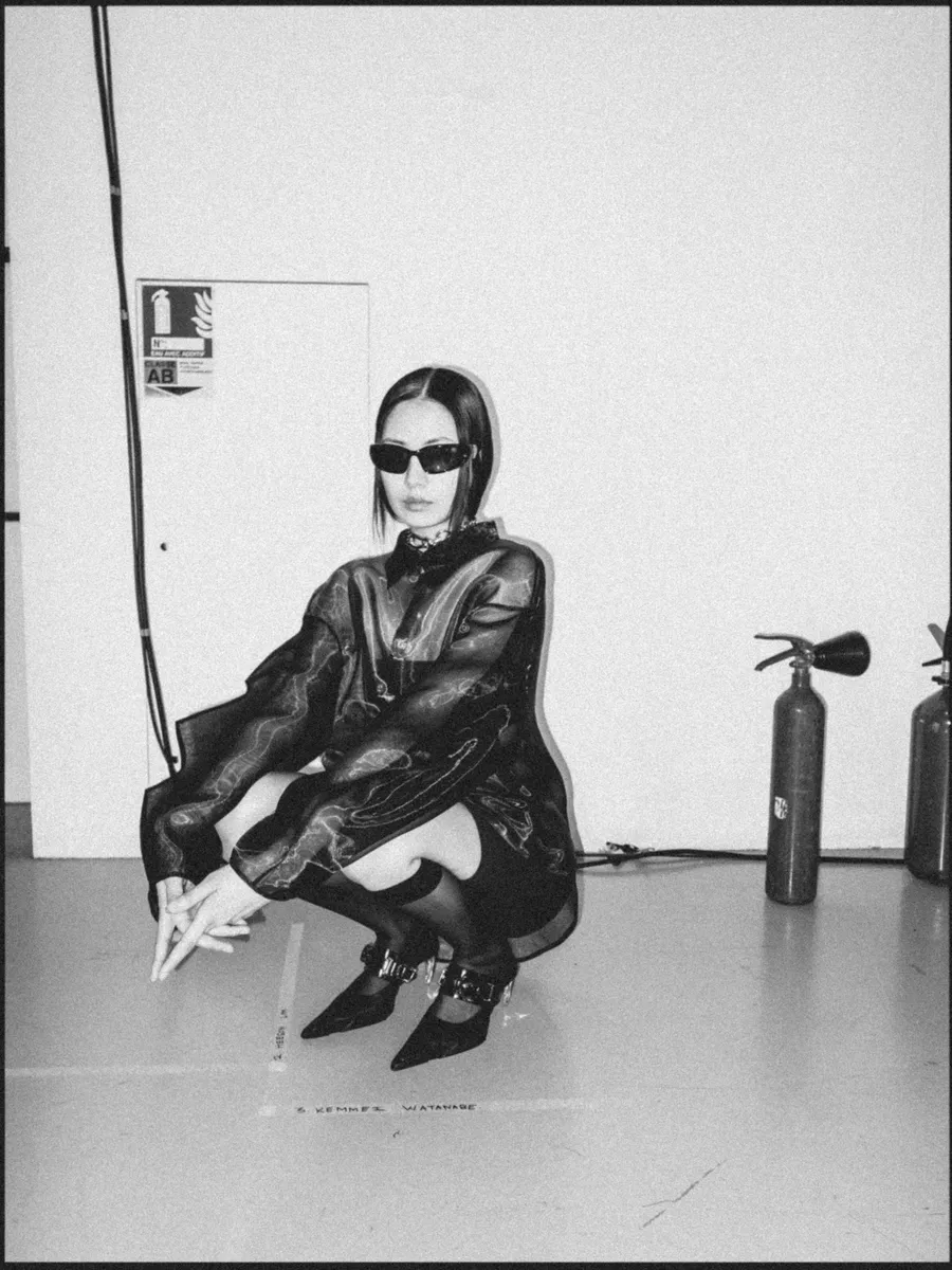 Femme accroupie en lunettes noires et veste ample imprimée, extincteurs au sol, noir et blanc