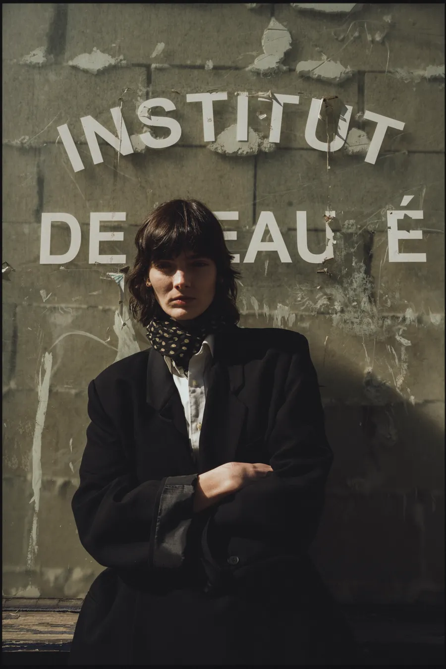 Femme bras croisés en veste noire oversize et foulard à pois, devant un mur portant l'inscription « Institut de beauté »