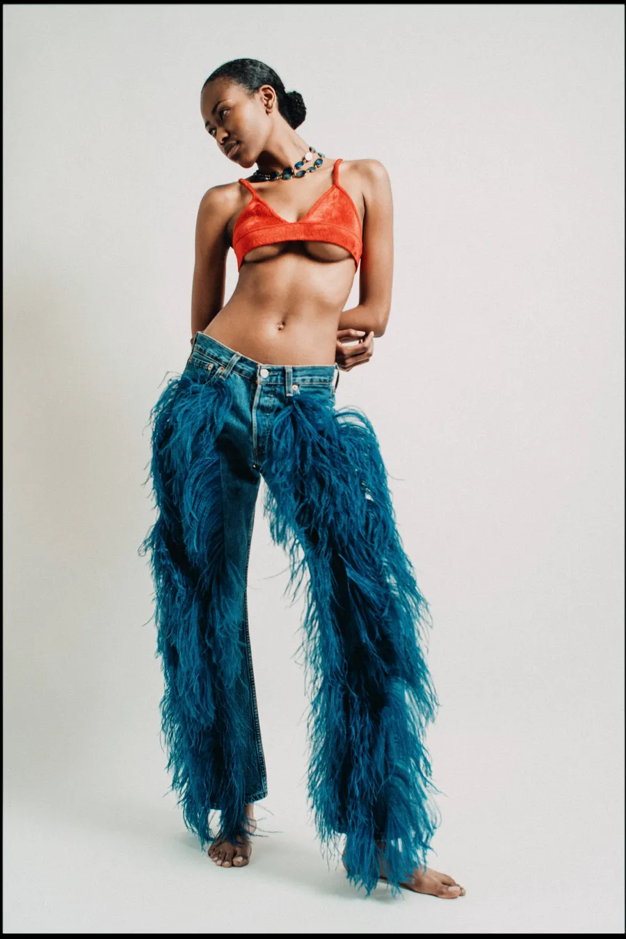 Mannequin pieds nus en brassière rouge et jean orné de plumes bleues, fond de studio clair