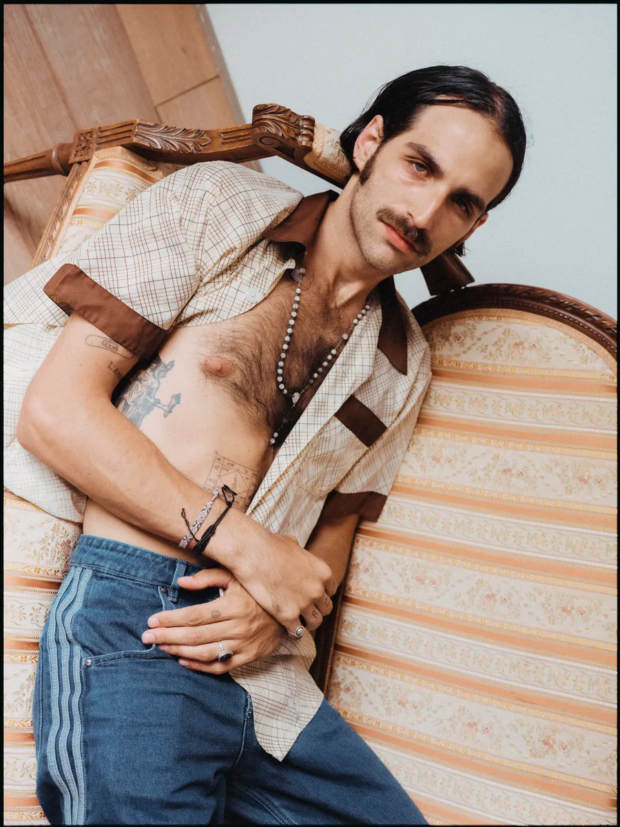 Homme moustachu allongé sur un fauteuil rayé, chemise ouverte, colliers et jean, torse visible