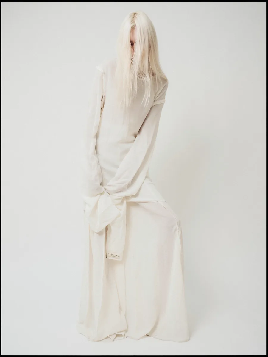 Silhouette en longue robe écrue à manches démesurées, visage masqué par de longs cheveux blonds, fond blanc