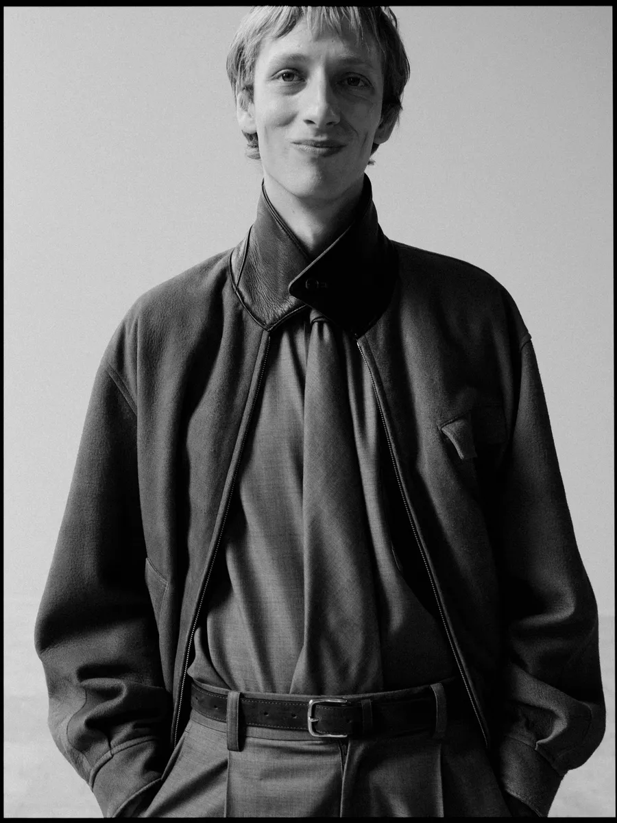 Homme souriant, mains dans les poches, blouson zippé à col de cuir sur chemise et ceinture, noir et blanc