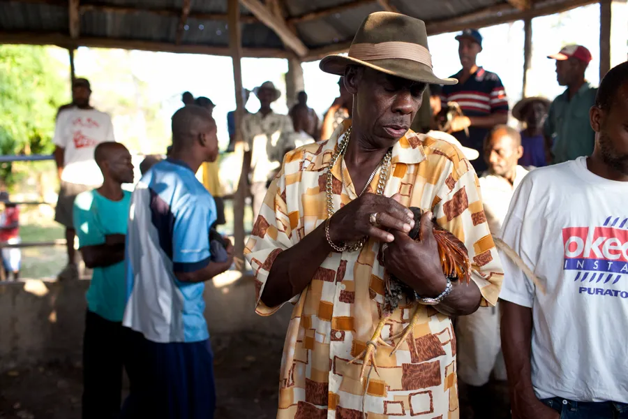 Homme au chapeau tenant un coq contre lui sous un hangar, groupe d'hommes autour
