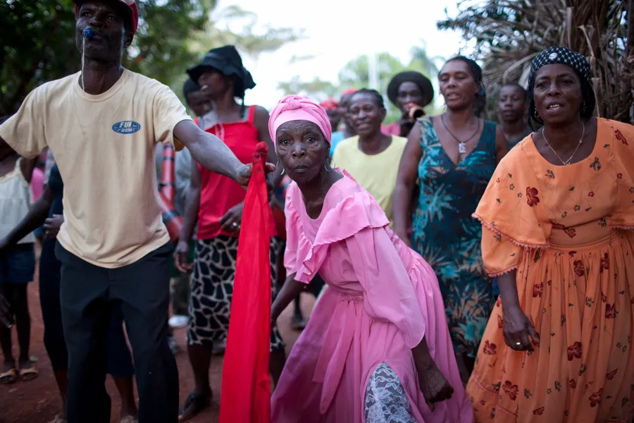 Femme en robe rose au milieu d'un cortège, homme tendant une étoffe rouge, chemin de terre