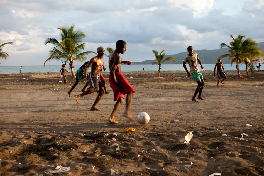 Jeunes hommes jouant au football pieds nus sur une plage de sable, cocotiers et mer derrière
