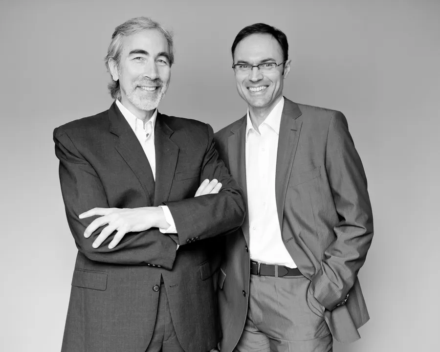 Deux hommes en costume souriant côte à côte devant un fond clair, noir et blanc