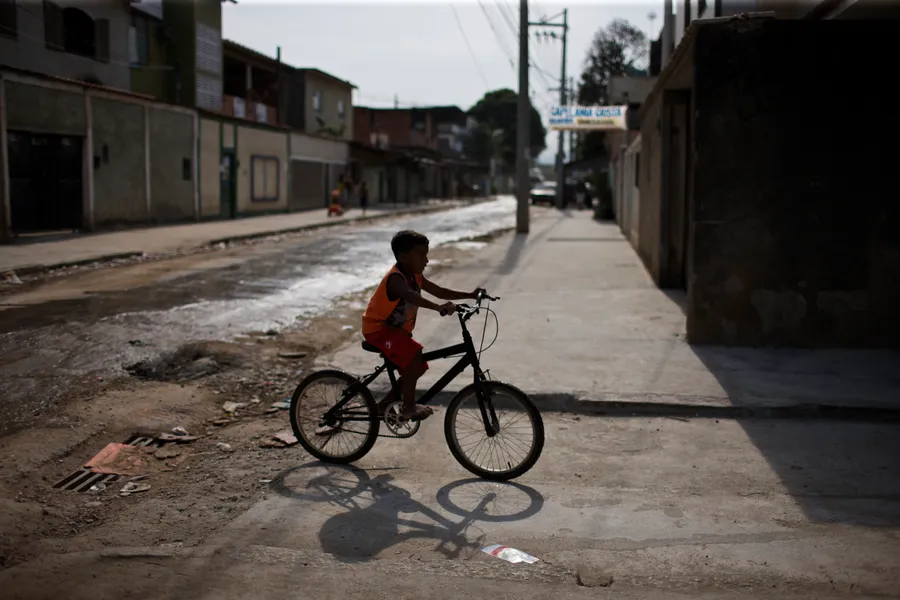 Garçon à vélo sur un trottoir en contre-jour, rue de favela et maisons basses
