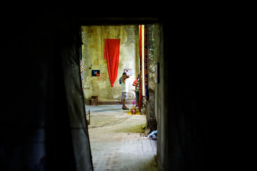 Vue par une porte sombre sur une cour où un homme se tient devant une tenture rouge