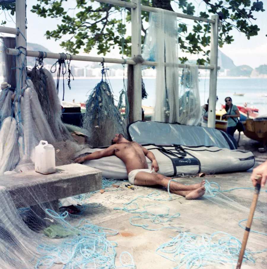 Homme endormi torse nu parmi des filets de pêche et des cordages bleus, plage et ville derrière