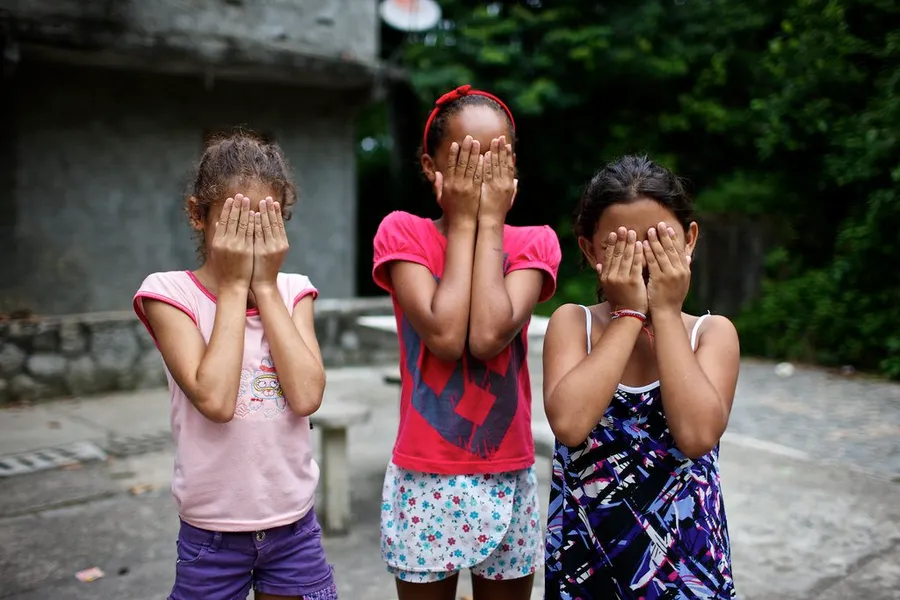Trois fillettes debout côte à côte, mains devant le visage, cour bétonnée et végétation