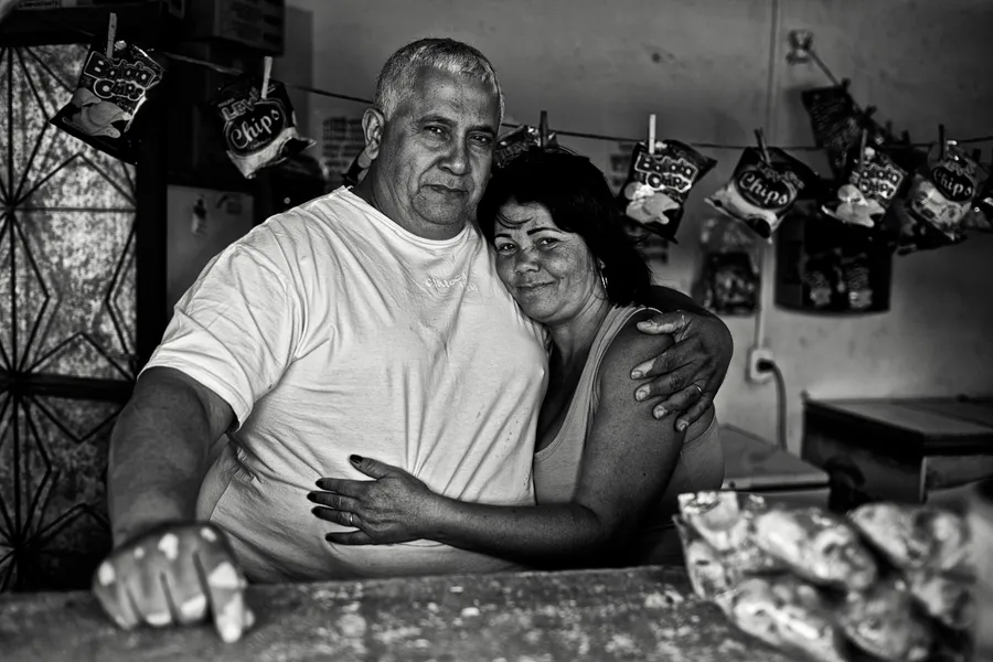 Couple enlacé derrière le comptoir d'une petite épicerie, sachets de chips suspendus, noir et blanc