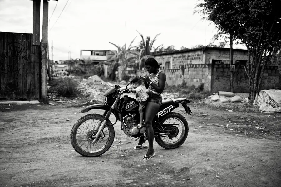 Femme debout près d'une moto avec un enfant assis sur la selle, terrain vague, noir et blanc