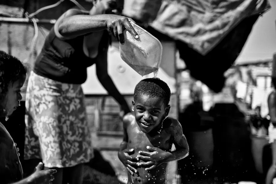 Femme versant l'eau d'un bidon sur la tête d'un enfant qui rit, linge suspendu, noir et blanc