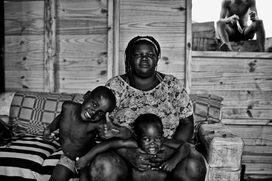 Femme assise sur un canapé avec deux jeunes enfants, cloison de bois clair derrière, noir et blanc