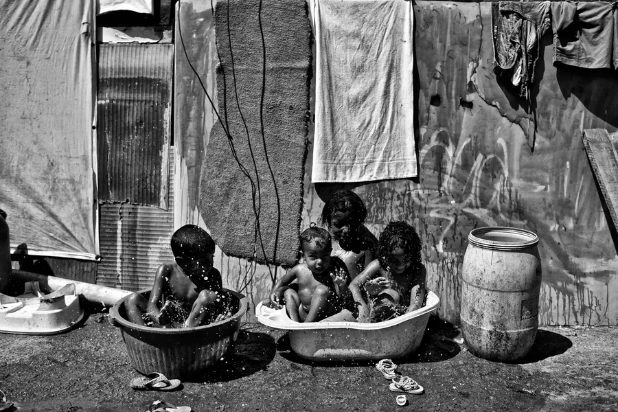 Quatre enfants prenant leur bain dans des bassines devant un mur de tôle, noir et blanc