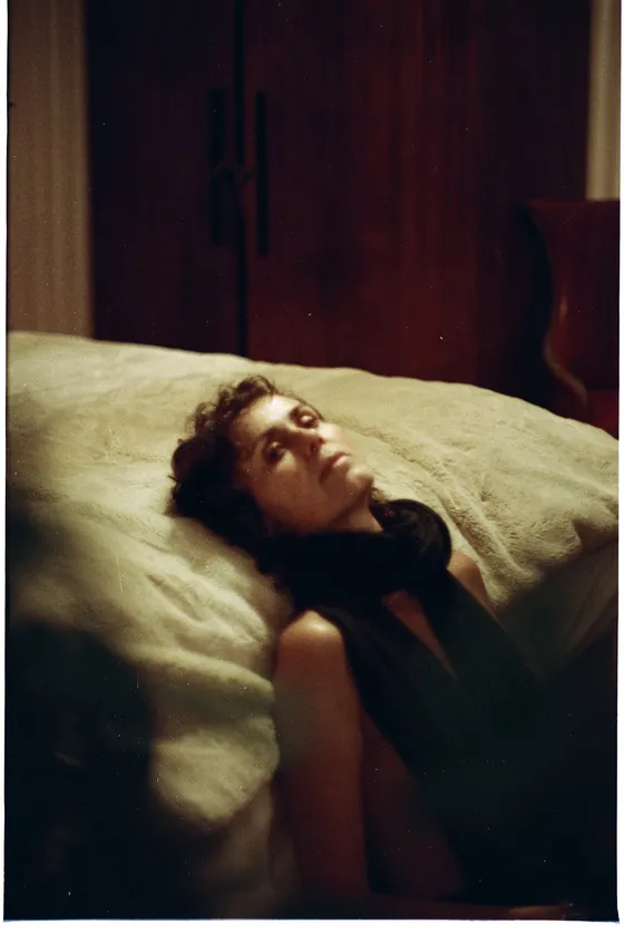 Femme aux cheveux bouclés allongée sur un lit, tête renversée, robe noire, chambre à la lumière tamisée